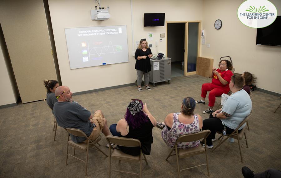 People sitting in a semicircle of chairs. A woman is presenting in the front of the room