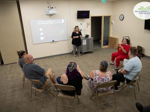 People sitting in a semicircle of chairs. A woman is presenting in the front of the room